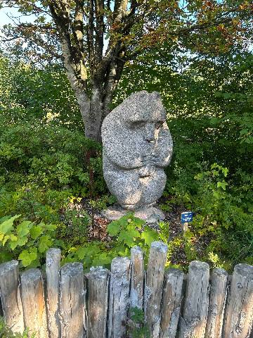a stone sculpture of a humanoid figure in a garden with a wooden fence